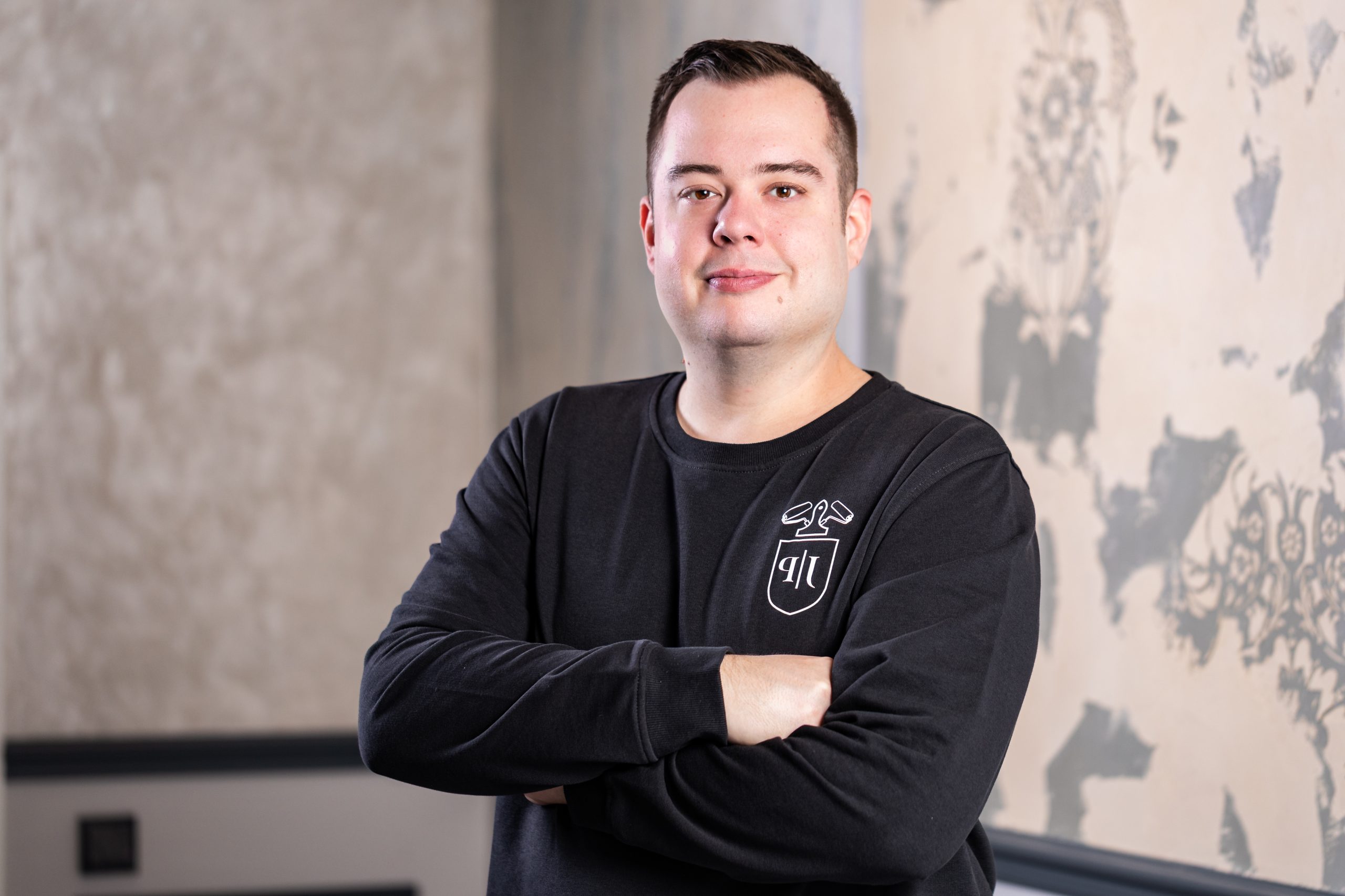 Michael Dettmann steht mit verschränkten Armen vor einer gemusterten Wand und blickt freundlich in die Kamera. Er trägt dunkles Sweatshirt mit Logo auf der Brust.